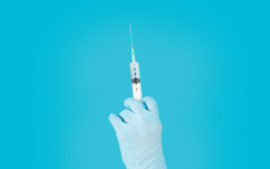 A gloved hand holds a syringe filled with liquid against a bright blue backdrop. The syringe is...