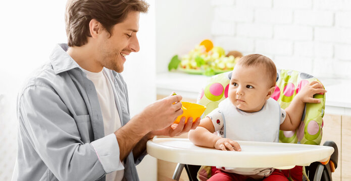 A father is feeding his baby in a high chair in a kitchen. The baby is wearing a bib and sitting in a brightly colored high chair. The father is smiling and looking at his baby.