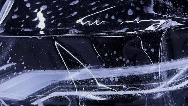 Employees of a car detailing studio apply clear protective PPF film on the hood of a black car. The video showcases the meticulous process ensuring high-quality protection and finish.
