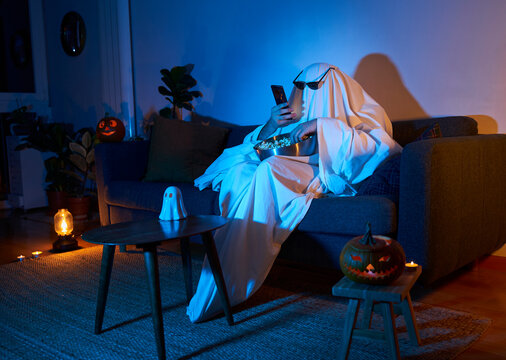 Halloween scary movie night. Person in ghost costume with sunglasses and pumpkin decor texting and eating popcorn while watching tv in sofa