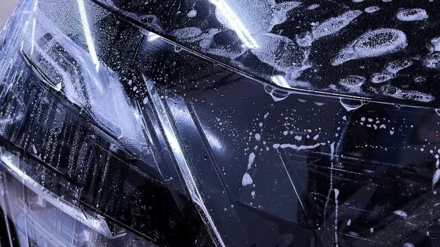 Employees of a car detailing studio apply clear protective PPF film on the hood of a black car. The video showcases the meticulous process ensuring high-quality protection and finish.