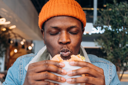 African American man savoring delicious burger in cozy cafe, capturing the joy of eating. Hungry guy enjoys biting into burger, eating fast food, unhealthy food.