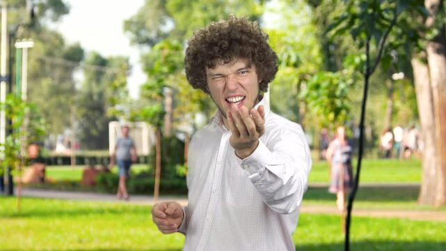Portrait of a young guy with curly hair gives you an air kiss outdoors. Public summer park in the background.