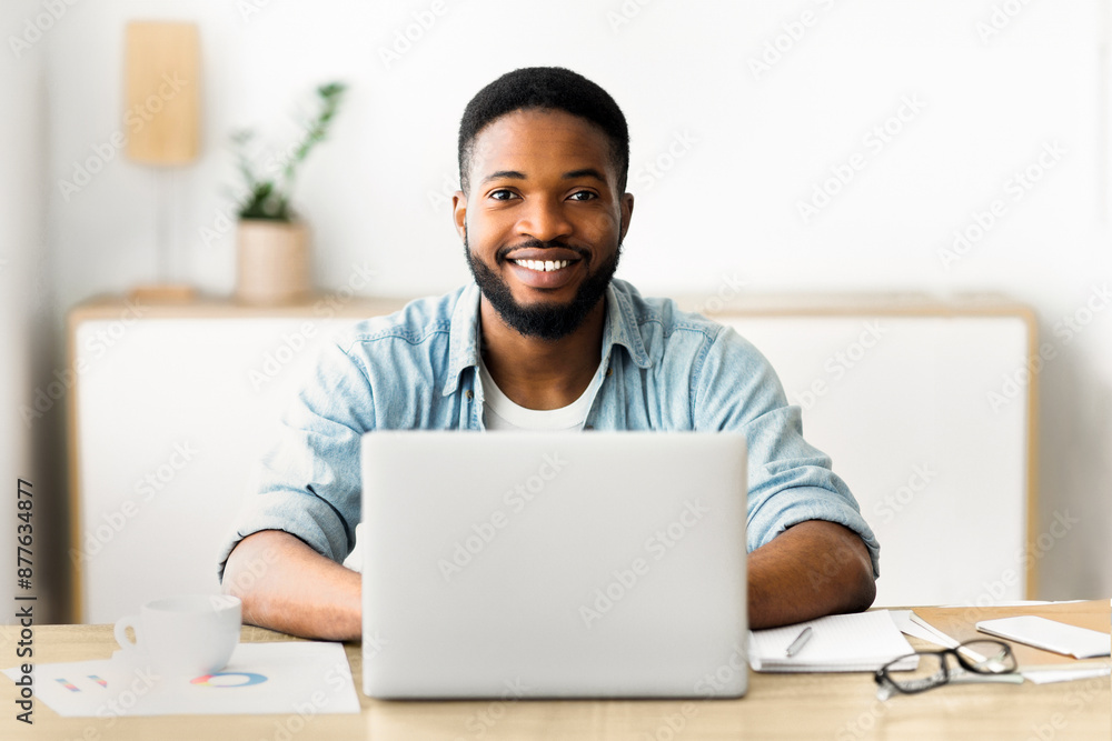 Poster a man with a beard smiles as he works on a laptop at a desk.