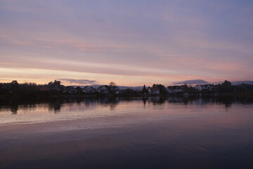 Fototapeta premium A serene lakeside view at dusk, with pastel hues of pink, purple, and blue painting the sky. The calm water reflects the charming houses lining the shore.