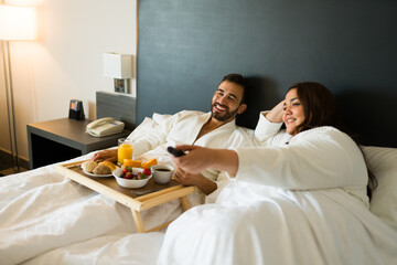Cheerful couple relaxing in bathrobes, having breakfast in bed and watching tv in their hotel room