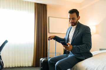 Content businessman sitting on a bed in a hotel room, smiling while using a smartphone and holding a suitcase