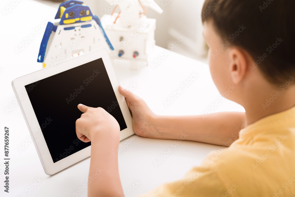 Poster A young boy, sitting at a table, uses a tablet with his right hand and points at the screen. The screen is black. There is a partially constructed robot in the background.