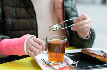  woman with a pink fiberglass cast on her right hand sits in a cafe at a table holding a mug of hot tea in her hand, brewing tea with a teapot,