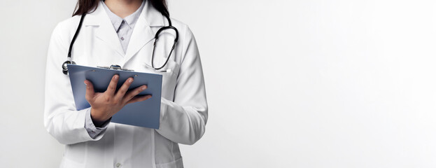 A doctor wearing a white coat and stethoscope holds a clipboard in front of a white background.