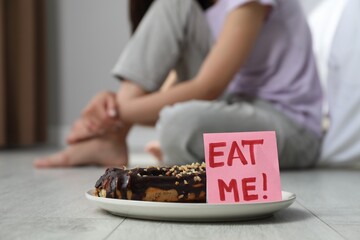Eating disorder. Woman sitting near note with words Eat Me and eclair on floor indoors, selective...