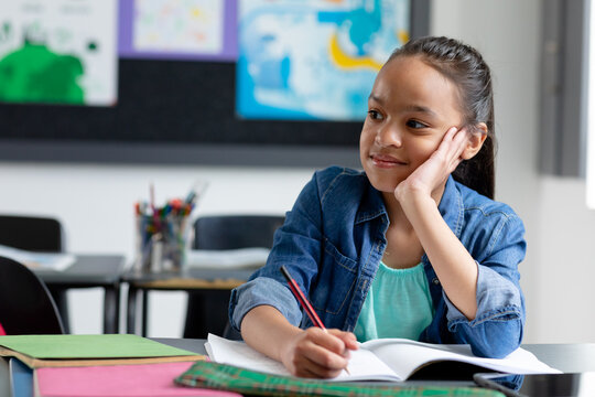 Bored biracial schoolgirl sitting at desk in class, leaning on hand, looking away, with copy space