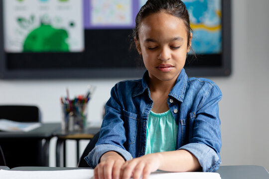 Blind biracial schoolgirl sitting at desk in class reading braille, with copy space - Powered by Adobe