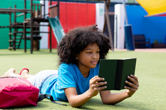 Smiling biracial schoolgirl lying in school playground reading book, with copy space