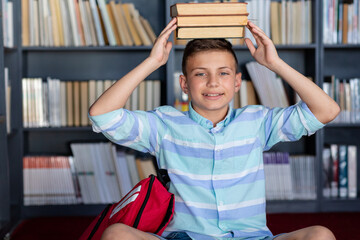 Smiling caucasian schoolboy with books on his head in library, copy space