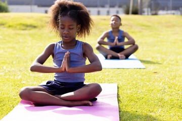 Happy african american schoolchildren doing yoga and meditating on field at school