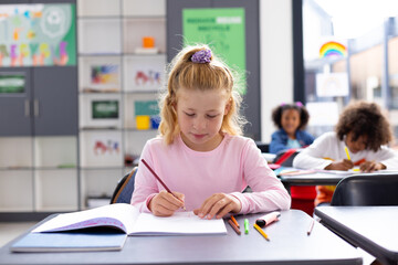 Fototapeta premium Happy caucasian schoolgirl sitting at desk and writing in notebook in school classroom