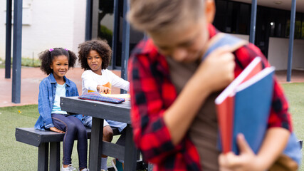 Sad caucasian schoolboy holding notebooks with diverse schoolchildren in background