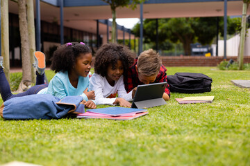 Happy diverse schoolchildren using tablet and sitting on grass at school