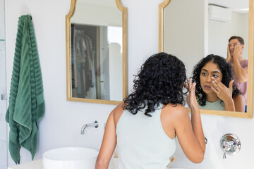Applying makeup, woman looking in mirror while man washing face in bathroom