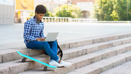 This image shows a teen black guy sitting on a set of stone steps in a city environment. He is using a laptop and has a skateboard beside him, copy space © Prostock-studio