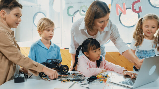 Smart teacher and diverse student looking at laptop on table with car model placed. Caucasian mentor inspect, explain, teaching engineering code or prompt in STEM technology classroom. Erudition.