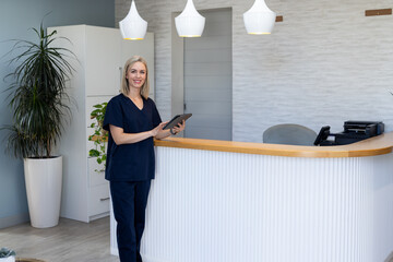 Standing at reception desk, woman in scrubs holding tablet in medical clinic, copy space