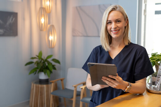 Smiling woman in scrubs holding tablet, standing in medical clinic reception area, copy space - Powered by Adobe
