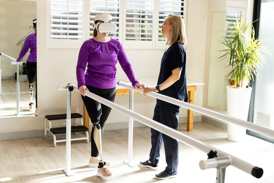 Woman in prosthetic leg walking using VR headset with therapist on parallel bars - Powered by Adobe