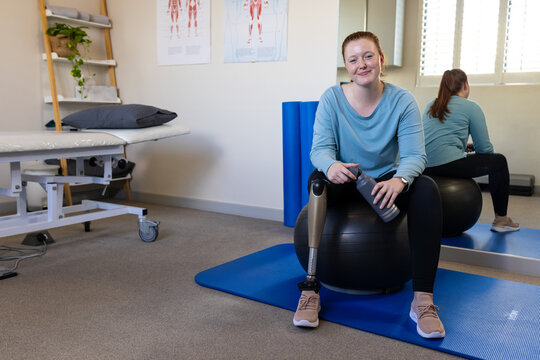 Sitting on exercise ball, woman with prosthetic leg holding water bottle in medical clinic, copy spa - Powered by Adobe