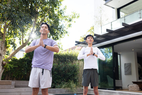 Practicing yoga, two men standing in prayer pose outdoors at home