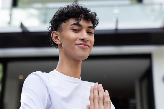 Smiling young man with hands in prayer pose, standing outside modern home