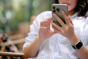 Close-up woman using a smartphone or mobile phone while sitting on wooden chair with green bokeh background.