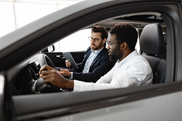 Car dealership, sales, and customer service. Caucasian car dealer sit inside vehicle with African...