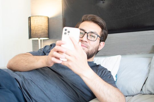 Hombre apuesto con anteojos relajado en su habitaci&oacute;n. Joven apuesto usando su tel&eacute;fono celular