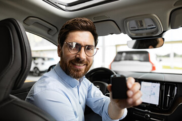 Happy expression, successful purchase or sale of automobile. Smiling businessman holding car key inside modern vehicle, sitting in dealership. Professional environment, modern technology.