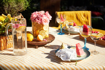 A bright summer garden setting in a yellow palette, with a yellow tablecloth, a vase of lemons with garden roses and watermelons