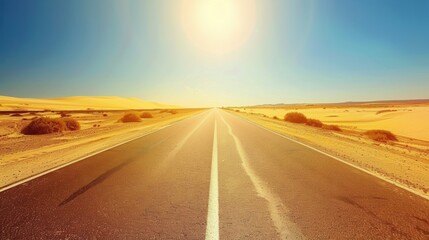 Naklejka premium A highway with a dashed white line down the center, stretching into the horizon through a desert landscape under a blazing hot sun. The road is flanked by sand dunes and sparse desert vegetation, with