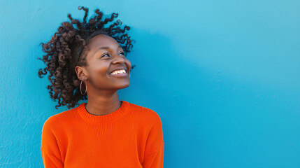 Joyful woman with curly hair smiling brightly against a vibrant blue background, wearing an orange sweater.