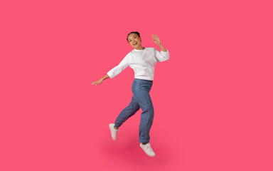 Asian young woman jumps and smiles in front of a bright pink background, her arms outstretched and her white shirt billowing in the air.