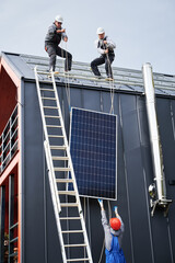 Men roofers installing solar panel system on roof of house. Technicians in helmets lifting up...