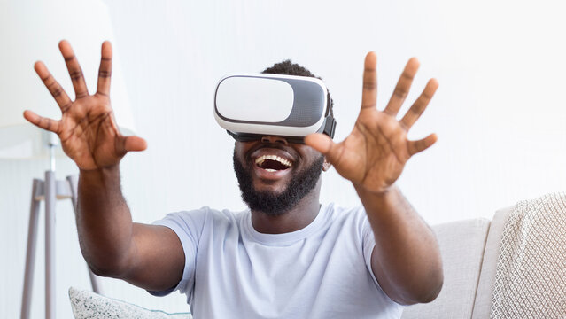 African American man wearing a VR headset is seated on a couch, both hands outstretched, as he laughs in delight at something in the virtual world.