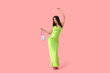 Beautiful young woman in stylish green dress with handbag dancing on pink background