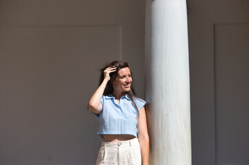 Fototapeta premium young and beautiful latin woman is leaning on a white marble column and touching her hair with her hands. The woman is happy and smiling on the sunny day in the city of Seville, Spain.