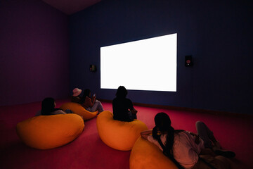 White cinema screen mockup with silhouettes of spectators setting on beanbags in colorful room