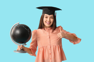 Young female graduate with globe showing thumb-up on blue background