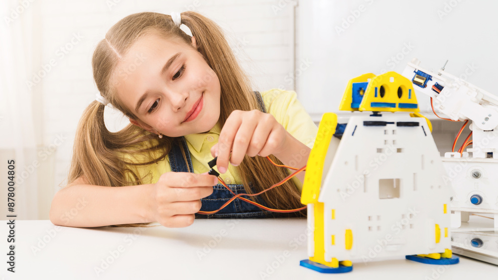 Poster a young girl with pigtails smiles as she builds a robot kit at a white table. she is holding a wire 