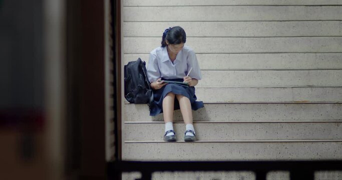 Thai or Asian high school girls in cute white school uniforms, using tablets and pens for practicing reading and revising knowledge, sitting on school stairs in the quiet evening after school.