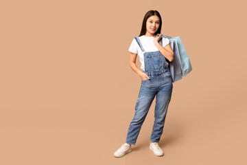 Naklejka premium Fashionable young Asian woman in stylish denim overalls with shopping bags on brown background