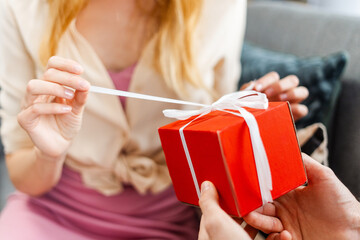 Woman untying white ribbon of red gift box being held by man, selective focus